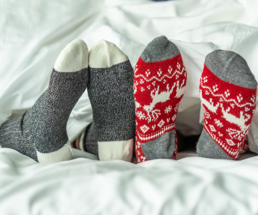 Stopy dwóch osób w grubych skarpetkach – jedna para w szaro-białe pasy, druga w czerwono-białe wzory z reniferami – wystające spod białej pościeli, co jest zaskakującym sposobem na bezsenność, o którym przeczytasz na Poradnik Zdrowie.