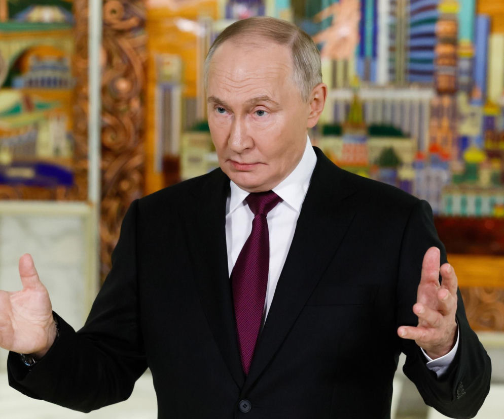 Prezydent Rosji Władimir Putin w czarnym garniturze i purpurowym krawacie, z otwartymi dłońmi, gestykuluje podczas wystąpienia na tle kolorowej, abstrakcyjnej dekoracji. Temat reparacji od Rosji dla Polski poruszono na Super Biznes.