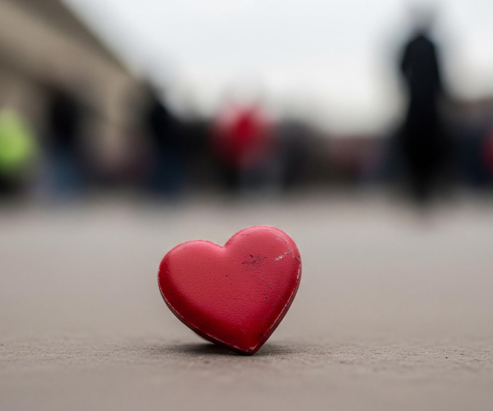 Czerwone serce, z lekkimi zadrapaniami i przetarciami na powierzchni, leży na jasnoszarej, ziarnistej nawierzchni, która rozciąga się od dołu kadru do jego środka. Obiekt jest ustawiony centralnie, skierowany lekko w prawo, z lewą krawędzią nieco wyżej niż prawa. Tło jest całkowicie rozmyte, ukazując niewyraźne kształty ludzi i innych obiektów w różnych kolorach, w tym fragmenty czerni, bieli, jaskrawej zieleni i czerwieni. Górna część tła jest jaśniejsza, przechodząca w biel ku górze.