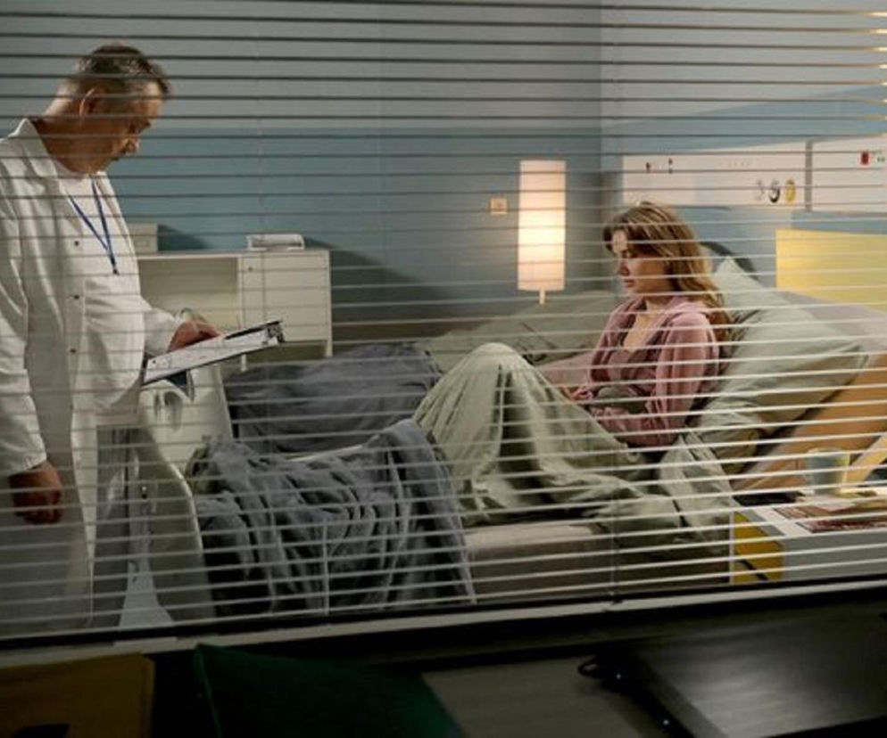 Lekarz Wojciech Billip w białym kitlu, z kartką w ręku, rozmawia z Franką (Dominika Kachlik) leżącą na łóżku szpitalnym w różowej bluzce, przykrytą kocem. Panika Franki przed operacją tętniakowo poszerzonej aorty w M jak miłość. Więcej o tym przeczytasz na Super Seriale.