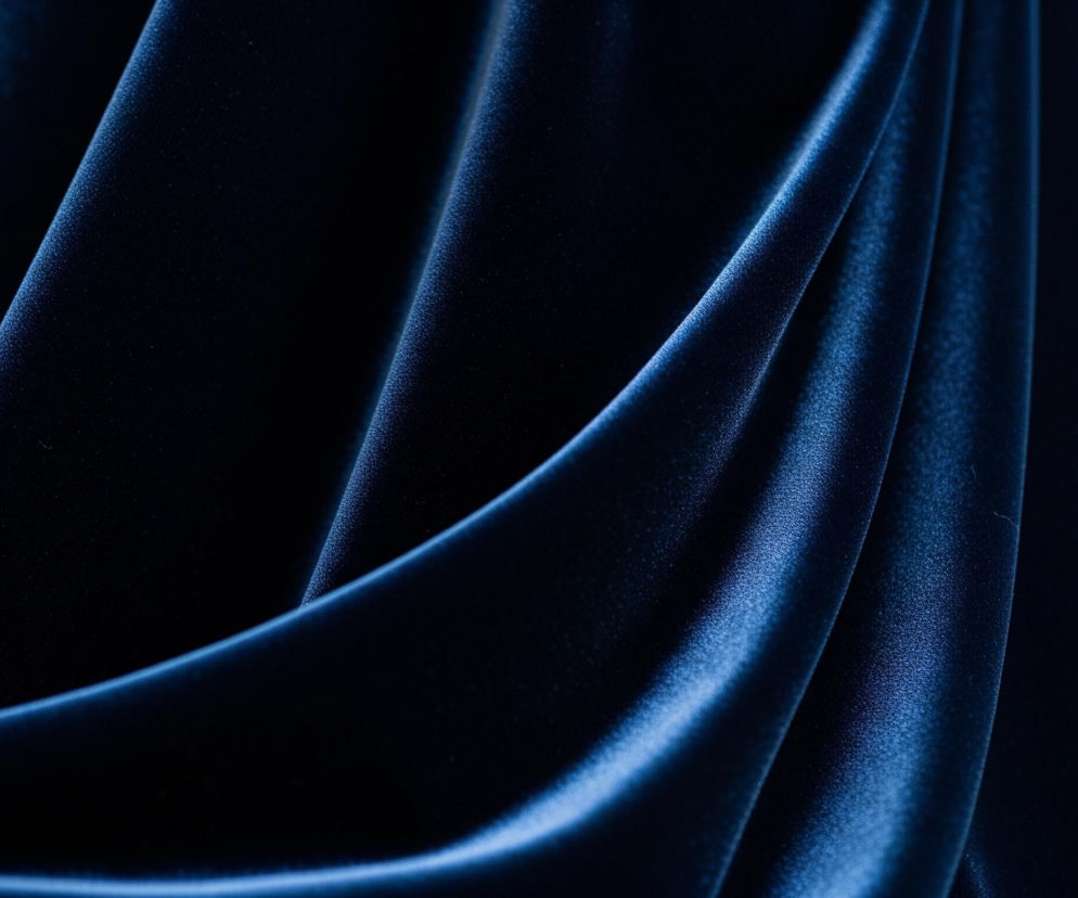 Centralny element obrazu to intensywnie niebieska, aksamitna tkanina, z wyraźnymi fałdami i draperiami. Faktura materiału jest miękka i błyszcząca, z mniejszymi i większymi zagięciami, które tworzą dynamiczne cienie i rozjaśnienia. Światło pada z lewej strony, podkreślając puszystość aksamitu i różnice w odcieniach niebieskiego – od głębokiego granatu w cieniach po jaśniejszy, niemal kobaltowy błękit na wypukłych częściach. W tle, po prawej stronie, widoczny jest rozmyty, jasnobrązowy obszar, a po lewej, w górnym rogu, niewyraźny fragment jasnego koloru.
