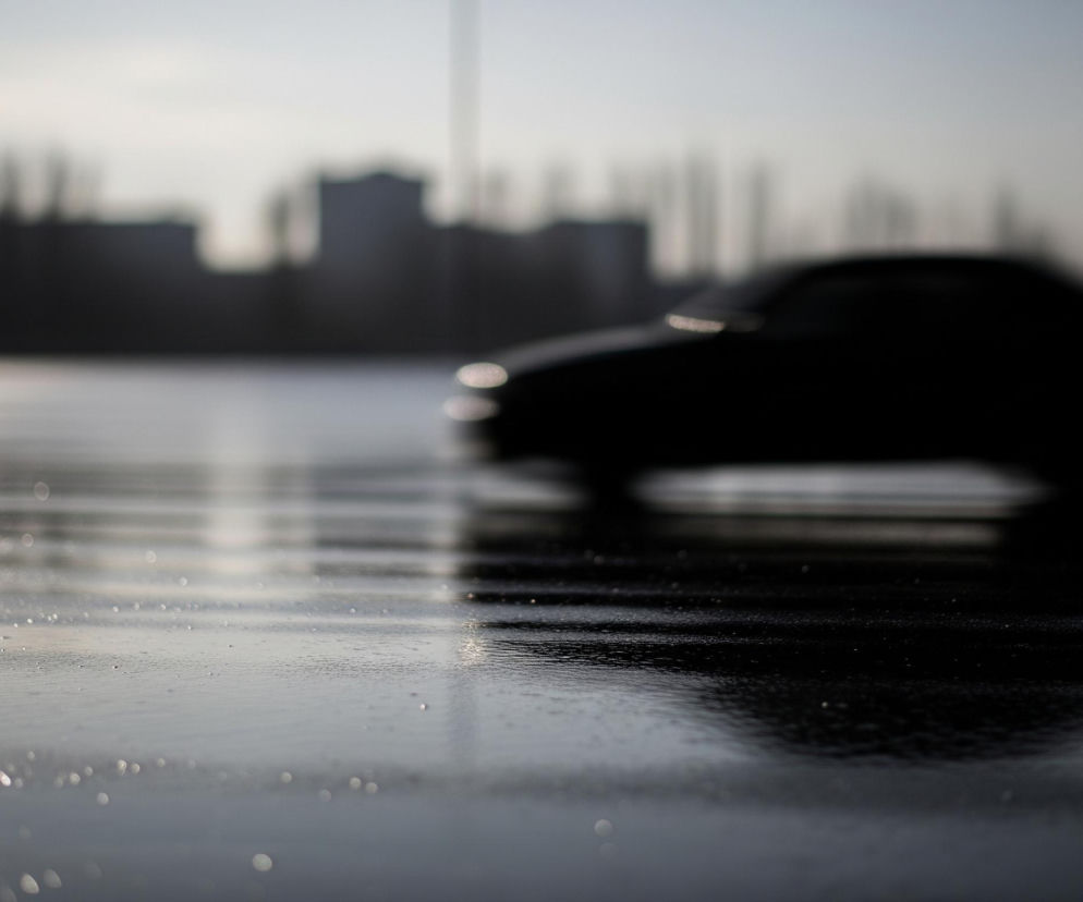 Pojazd o ciemnej karoserii, niewyraźny z powodu szybkiego ruchu, przemieszcza się po mokrej powierzchni, pozostawiając za sobą rozmyty ślad. Odbicia świateł na mokrej nawierzchni tworzą jasne plamy i linie, zlewając się z refleksami na przedzie samochodu. W tle widać ciemne, zamazane sylwetki budynków i drzew, a nad nimi jaśniejsze, szare niebo. Całość jest silnie rozmyta, z wyjątkiem detali na mokrej ziemi w dolnej części kadru.