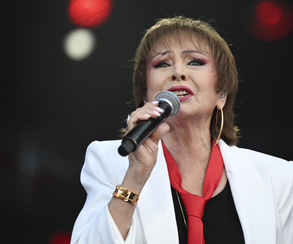 Izabela Trojanowska śpiewa na scenie, ubrana w białą marynarkę i czarną bluzkę z czerwonym krawatem. Wokalistka trzyma mikrofon i ma wyraźny makijaż. Artystka opowiedziała o swoim podejściu do Tańca z Gwiazdami, o czym przeczytasz więcej na naszym portalu.