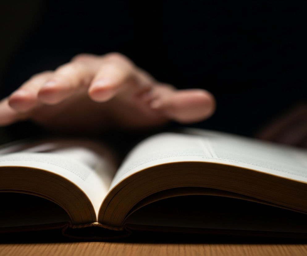 Otwarta książka z jasnymi, tekstowymi stronami spoczywa na drewnianym stole, zajmując centralną część kadru. Jej kartki są nieco pożółkłe na krawędziach, a grzbiet książki jest ciemnobrązowy, rzucając cień. Nad książką, w górnej części zdjęcia, widać niewyraźną, jasną dłoń skierowaną palcami w dół, zawieszoną nad stronami. Tło jest niemal całkowicie czarne, co sprawia, że światło skupia się wyłącznie na książce i częściowo na dłoni.
