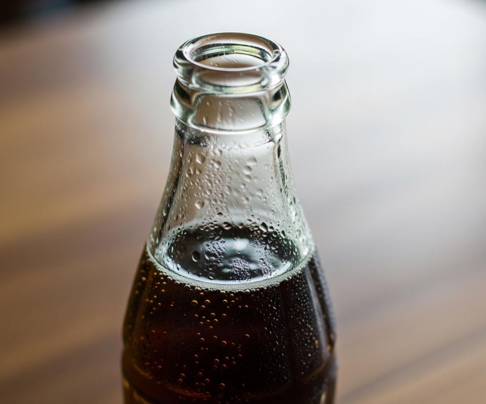 Przedstawiona jest zbliżenie na otwartą szklaną butelkę wypełnioną ciemnym płynem. Na zewnętrznej powierzchni butelki widoczne są liczne krople skondensowanej pary wodnej, a na powierzchni płynu unosi się pianka z bąbelkami. Butelka stoi na drewnianym, brązowym blacie, który jest rozmyty na dalszym planie.