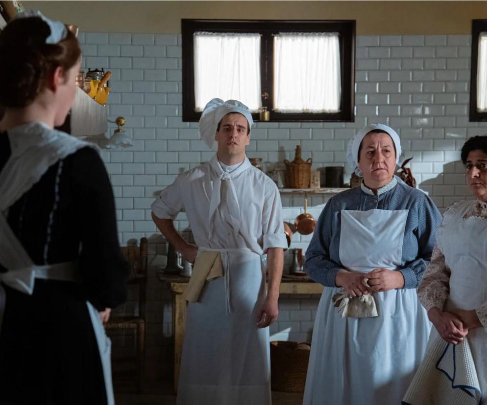 Czwórka służby pałacowej, w tym mężczyzna w stroju kucharza i trzy kobiety w tradycyjnych strojach służebnych, stoi w kuchni o białych kafelkowych ścianach. Kobieta na pierwszym planie stoi tyłem. Zdjęcie ilustruje odcinek serialu La Promesa, o którym możesz przeczytać na Super Seriale.