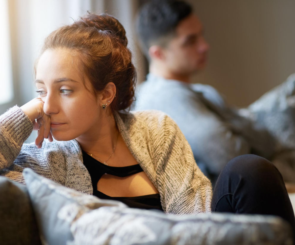 Kobieta w swetrze, zamyślona, z ręką pod brodą, siedząca na kanapie tyłem do mężczyzny, symbolizuje nostalgię i wycofanie. Poradnik Zdrowie wyjaśnia, jak nostalgia wpływa na psychikę i emocje.