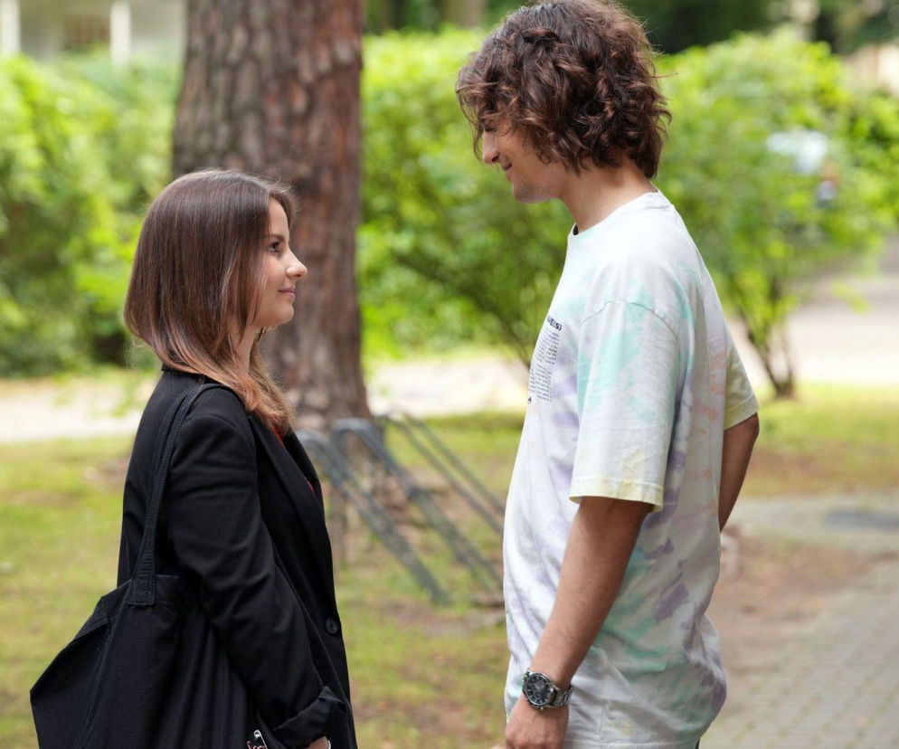 Ewa (Gabriela Ziembicka) i Romeo (Jacek Tyszkiewicz) patrzą na siebie z uśmiechem w parku, ona w czarnej marynarce i z torbą, on w T-shircie tie-dye. Romantyczna scena z serialu Barwy szczęścia dostępna na Super Seriale.