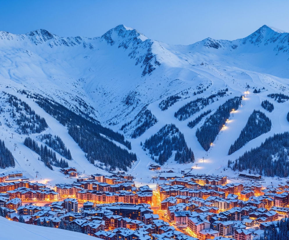 Śnieżne, poszarpane szczyty górskie dominują tło, zajmując górną połowę kadru pod błękitnym niebem. Stoki gór pokryte są grubą warstwą śniegu, przeplataną ciemnozielonymi kępami drzew iglastych i liniami wyciągów narciarskich, z których niektóre oświetlone są punktowym, ciepłym światłem. U podnóża gór rozciąga się oświetlone miasteczko, składające się z licznych budynków o spadzistych dachach, również pokrytych śniegiem, z których okien i wnętrz bije intensywne, pomarańczowe światło, kontrastujące z zimnym, niebieskim otoczeniem. Główna droga przecinająca miasteczko jest jasno oświetlona, tworząc smugę światła prowadzącą w głąb obrazu.