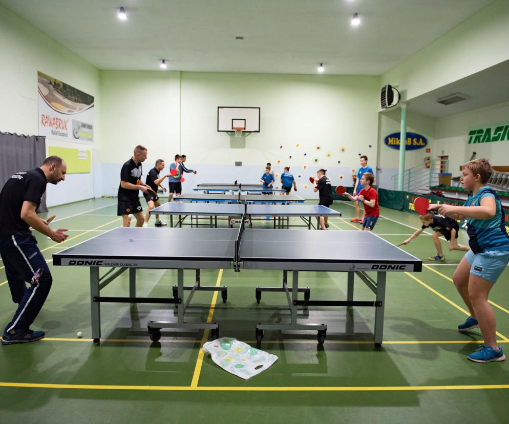 Dzieci i dorośli grający w tenisa stołowego na hali sportowej. O naborze do Akademii przeczytasz na Eska Ostrzeszów.