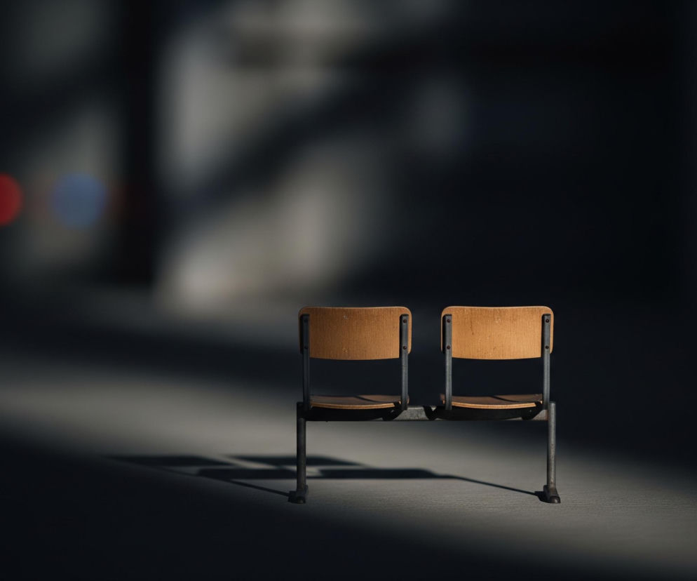 Na pierwszym planie widoczne są dwa puste krzesła z jasnobrązowymi, drewnianymi oparciami i siedzeniami, zamocowanymi na ciemnych, metalowych ramach. Krzesła stoją na jasnoszarej powierzchni, która rozciąga się w lewą stronę, częściowo oświetlona silnym, ciepłym światłem padającym od góry po lewej stronie. Cienie rzucane przez krzesła są długie i wyraźne, wskazując kierunek oświetlenia. Tło jest bardzo ciemne, niemal czarne, z delikatnymi, rozmytymi plamami światła i trzema mocno rozmytymi kolorowymi punktami w lewej górnej części: czerwonym, niebieskim i kolejnym czerwonym, które stapia się z niebieskim.