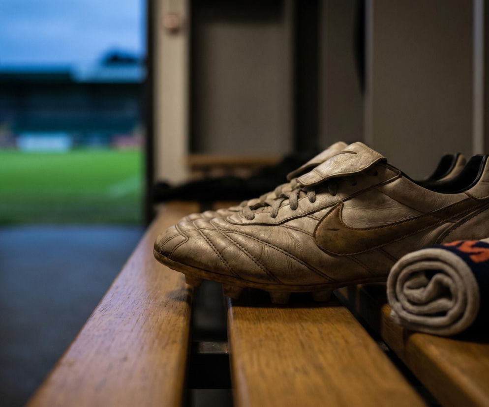 Stare, skórzane buty piłkarskie Nike Tiempo leżą na drewnianej ławce w szatni, obok zwiniętej skarpetki, symbolizując pożegnanie z klubem Lech Poznań i transfery w Ekstraklasie, o których przeczytasz na naszym portalu. W tle widać zamazane boisko.