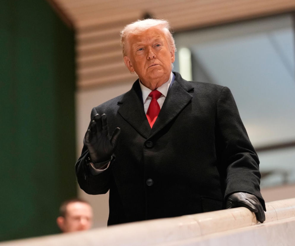 Donald Trump w czarnym płaszczu, białej koszuli i czerwonym krawacie, z dłonią uniesioną w geście, patrzy poważnie przed siebie. Tło jest rozmazane, sugerując wnętrze budynku. Zdjęcie ilustruje temat gróźb Trumpa wobec Europy, o czym więcej przeczytasz na Super Biznes.