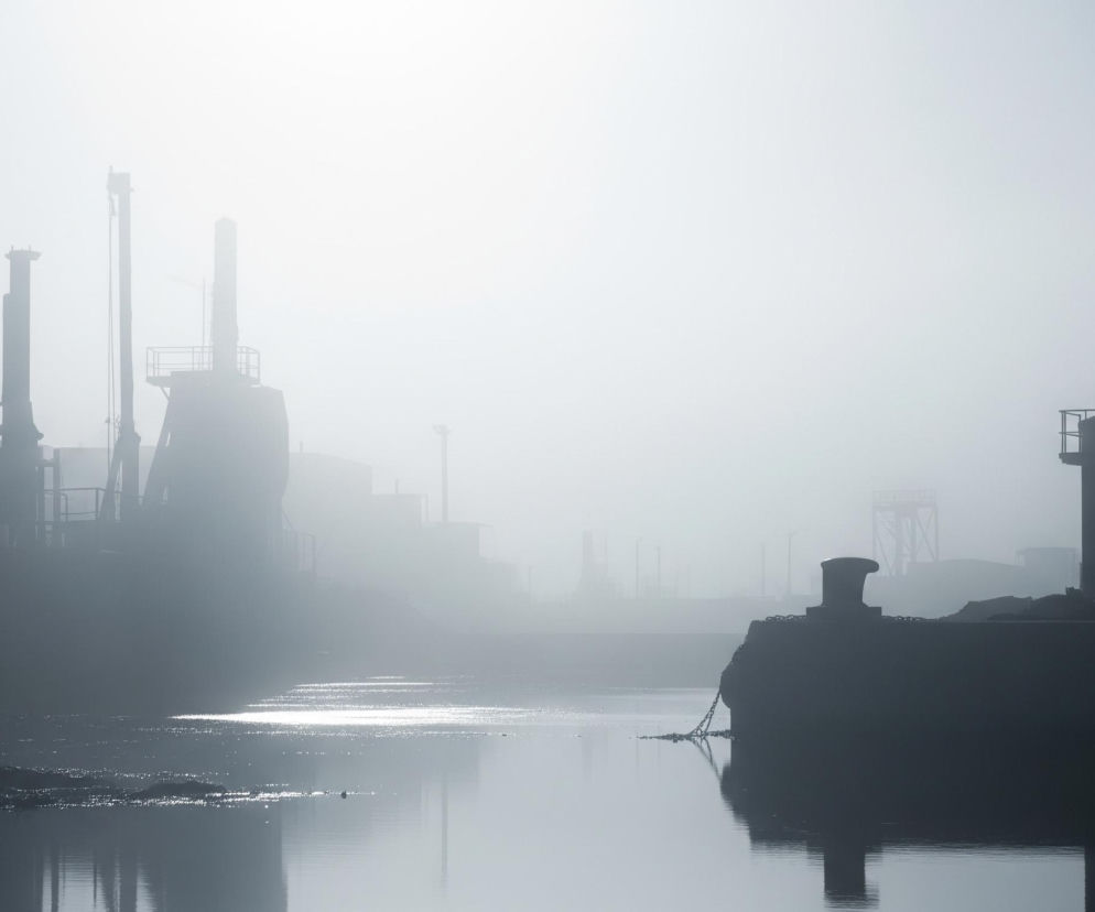Mocno zamglony pejzaż przedstawia industrialne nabrzeże, gdzie dominują ciemne, rozmyte sylwetki budynków i konstrukcji wznoszących się po obu stronach spokojnej tafli wody. Lewa strona ujawnia zarysy wysokich dźwigów portowych oraz budynków z kominami, spowitych gęstą mgłą, która stopniowo rozjaśnia się ku górze i centrum obrazu. Na pierwszym planie po prawej znajduje się ciemny, cylindryczny zbiornik, obok którego widoczny jest masywny poler, do którego przytwierdzony jest łańcuch delikatnie opadający w wodę, tworząc lustrzane odbicie. Pośrodku obrazu, na wodzie, widoczne są jasne, rozproszone refleksy światła, które delikatnie falują.