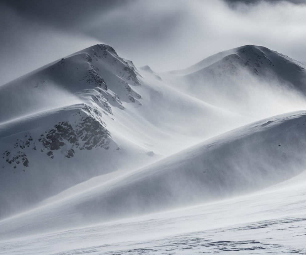 Widoczne są pokryte śniegiem, skaliste szczyty górskie, częściowo zasłonięte przez zamieć śnieżną. Ciemne, poszarpane skały przebijają się przez biały śnieg w lewej części kadru. Na pierwszym planie i w tle dominują zaśnieżone grzbiety, tworząc faliste kształty i ostre krawędzie. Chmury na niebie są ciemne po lewej stronie i jaśniejsze po prawej, zaś pędzący śnieg i wiatr zamazują granice między górami a niebem, nadając scenie dynamiczny, monochromatyczny charakter.