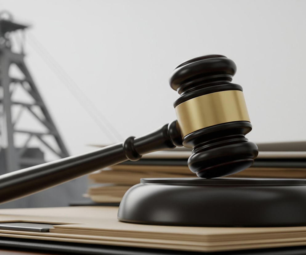 W centrum obrazu znajduje się czarny drewniany młotek sędziowski z błyszczącym, złotym pierścieniem na główce, leżący na czarnej, zaokrąglonej podstawce. Poniżej stosu dokumentów w odcieniach beżu i czerni, ułożonych na drewnianym blacie. W tle widoczna jest rozmyta, czarno-biała konstrukcja przypominająca wieżę szybową kopalni. Tło jest jasne, niemal białe, co kontrastuje z ciemnymi elementami na pierwszym planie.