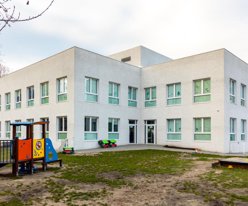 Zmodernizowany budynek Żłobka nr 31 w Warszawie, wyróżniony w konkursie Życie w Architekturze, z jasną elewacją i zielonymi oknami, otoczony placem zabaw z zabawkami. Obiekt prezentuje zrównoważoną adaptację istniejących struktur, o czym przeczytasz więcej na Architektura Murator Plus.