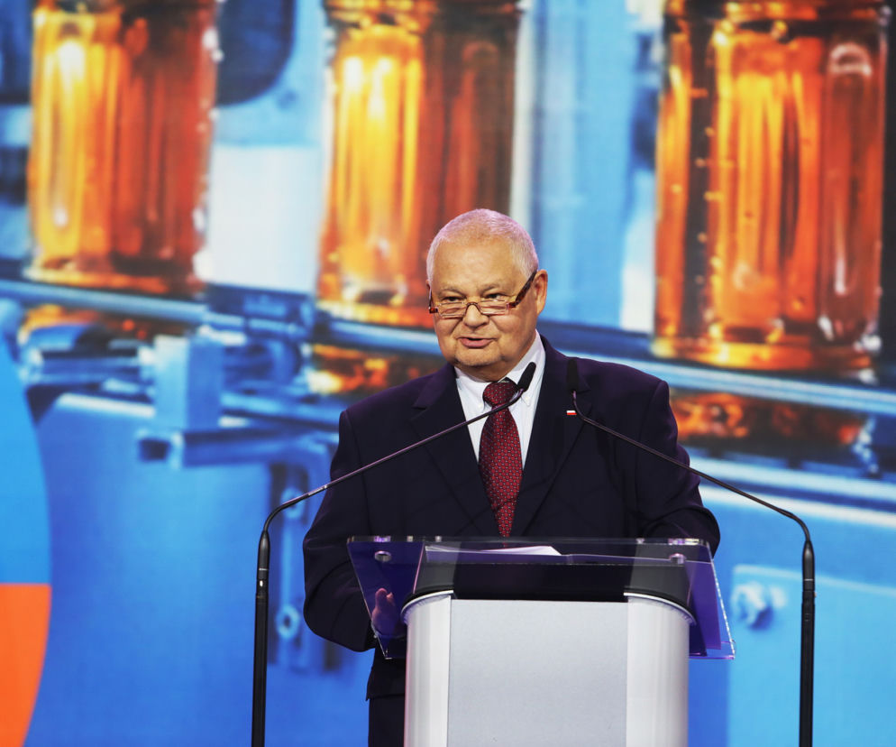 Prezes NBP Adam Glapiński w garniturze i krawacie, stojący za mównicą z dwoma mikrofonami. W tle widoczny ekran z abstrakcyjnym wzorem w kolorach pomarańczowym i niebieskim. Więcej informacji o konflikcie na Bliskim Wschodzie i stopach procentowych na Super Biznes.