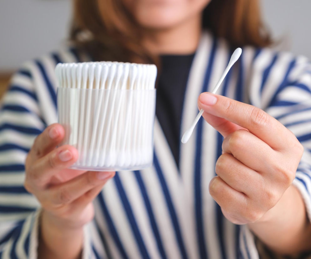 Kobieta trzyma w dłoniach opakowanie białych patyczków higienicznych i jeden patyczek osobno, skupiając wzrok na ryzyku ich używania do czyszczenia nosa. Na Poradnik Zdrowie znajdziesz więcej informacji o bezpiecznych metodach.