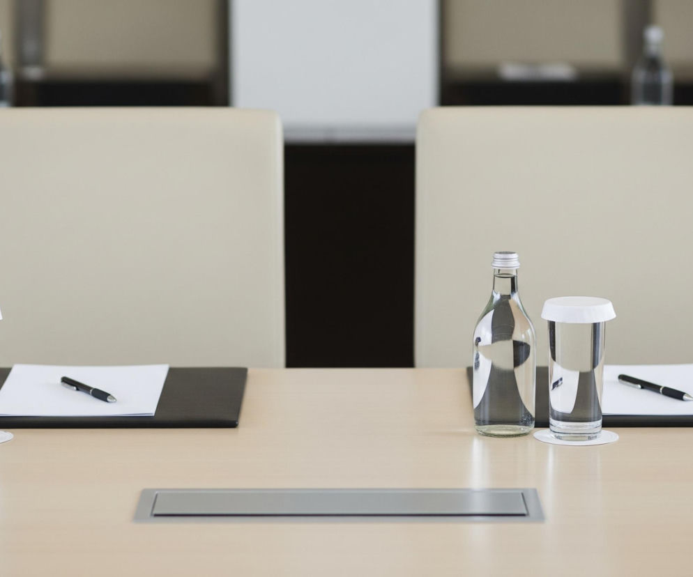 Na jasnobrązowym, polerowanym stole widoczne są dwa identyczne zestawy przedmiotów, symetrycznie rozmieszczone. Każdy zestaw składa się ze szklanej butelki z wodą, szklanki z wodą i białą pokrywką, oraz czarnej podkładki z białą kartką papieru i czarnym długopisem. W tle znajdują się dwa beżowe krzesła z oparciami oraz niewyraźne zarysy innych mebli i butelek z wodą. W centralnej części stołu znajduje się srebrny prostokątny panel.