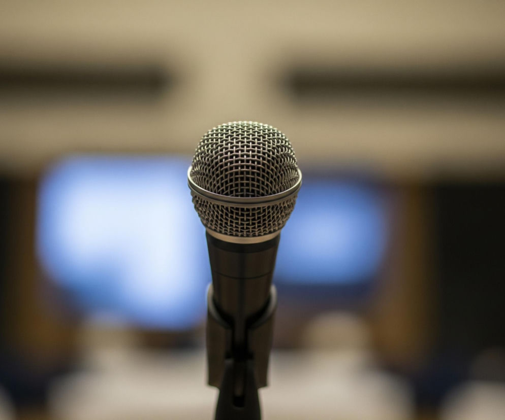 Na pierwszym planie, centralnie, widoczny jest czarny mikrofon z metalową, siatkową główką. Znajduje się on na czarnym stojaku, a jego górna część z siatki jest srebrzysta i odbija światło. Tło jest rozmyte, z jasnym, beżowym kolorem dominującym u góry i po bokach, oraz ciemniejszym, niebieskawym blaskiem w centrum, przypominającym ekran.