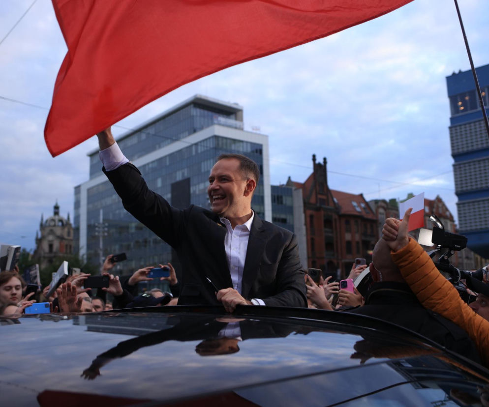 Uśmiechnięty Karol Nawrocki macha biało-czerwoną flagą z dachu samochodu. O sporach z rządem przeczytasz na SE Polityka.