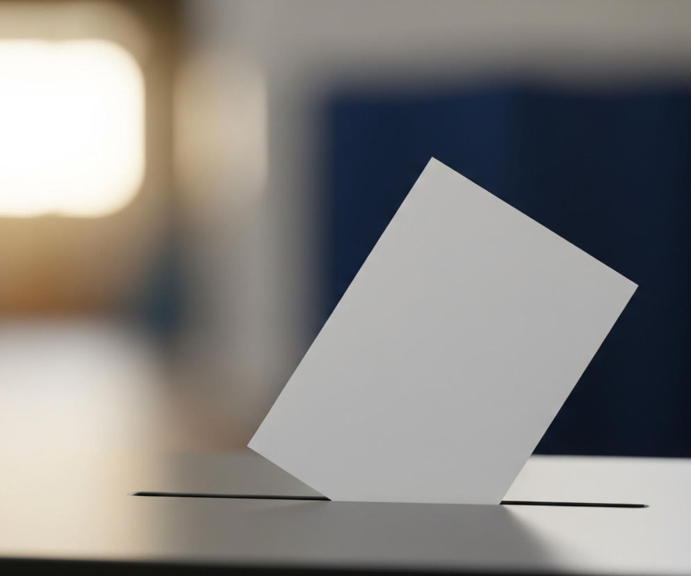 W centrum obrazu znajduje się biała, prostokątna kartka, częściowo włożona do wąskiej, poziomej szczeliny umieszczonej na górnej powierzchni ciemnoszarego obiektu. Kartka jest lekko przechylona w prawo. Tło jest rozmyte, z jasnym, rozświetlonym obszarem w kształcie zaokrąglonego prostokąta po lewej stronie i ciemniejszym, niebieskawym fragmentem po prawej. Całość utrzymana jest w stonowanej kolorystyce szarości, bieli i brązowo-niebieskich odcieniach, z miękkim oświetleniem.