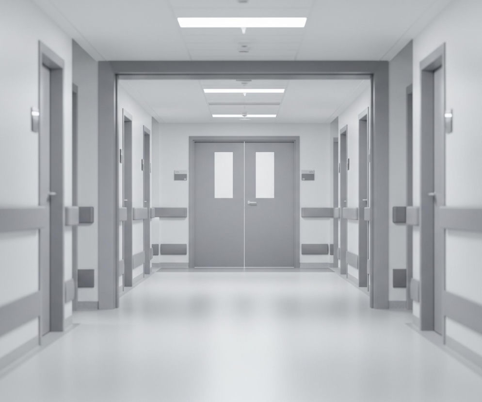 Długi, symetryczny korytarz szpitalny jest oświetlony od góry rzędami prostokątnych lamp. Ściany są jasnoszare, a podłoga jest gładka i odbija światło. Po obu stronach korytarza widoczne są szare drzwi, każde z prostokątnymi uchwytami i tabliczkami. Na wysokości około połowy wysokości drzwi biegną poziome, szare listwy ochronne, tworząc barierę wzdłuż ścian. Na końcu korytarza znajduje się para podwójnych, szarych drzwi z dwoma prostokątnymi oknami każde, prowadzące do kolejnego, podobnego korytarza.