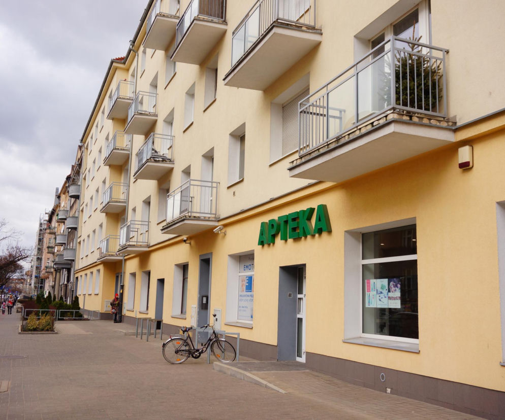 Apteka z zielonym szyldem i widocznym napisem „APTEKA” na tle kremowej elewacji budynku mieszkalnego, symbolizująca dostępność leków, która bywa zagrożona. Zobacz najnowsze informacje na ten temat na Poradnik Zdrowie.