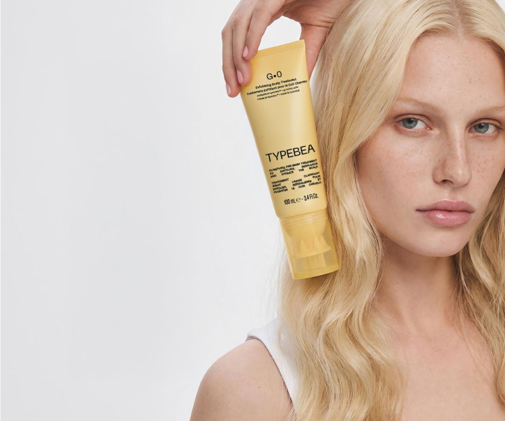 Kobieta o blond włosach trzyma żółtą tubkę peelingu do skóry głowy TYPEBEA. O nowościach w SE.