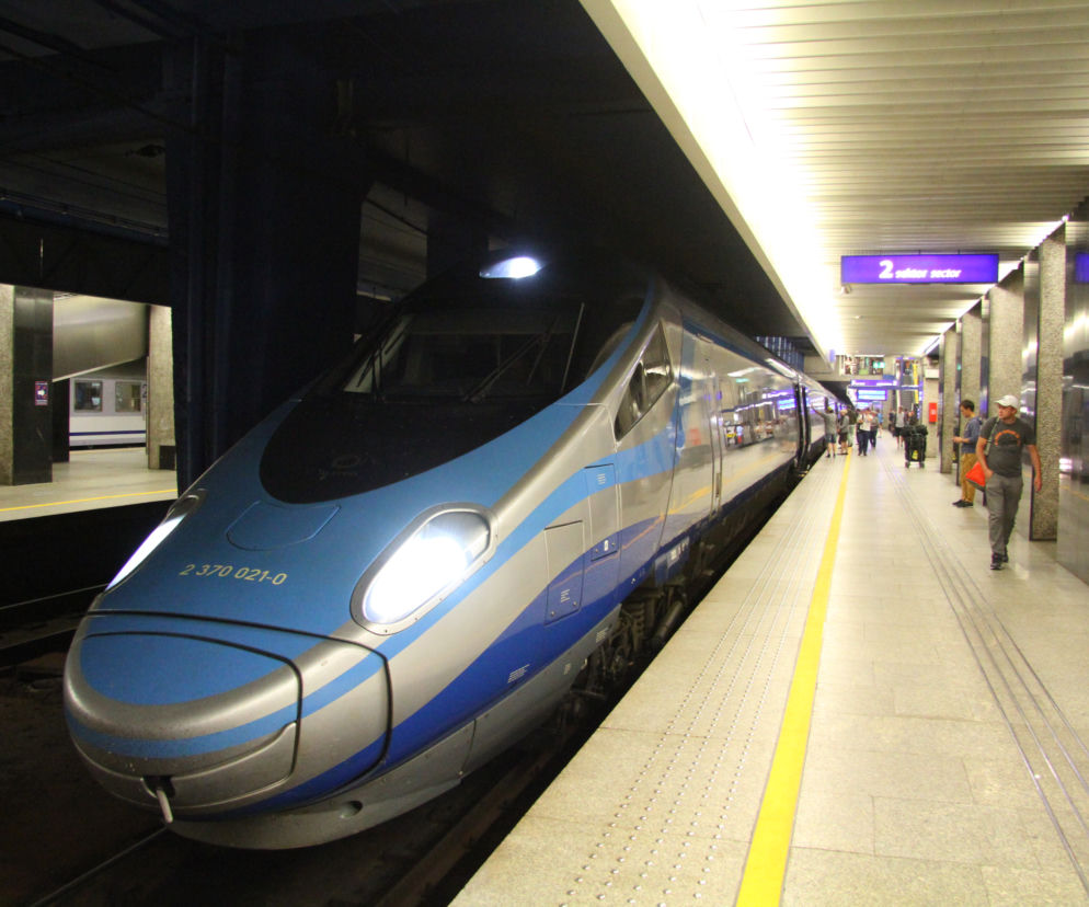 Nowoczesny pociąg Pendolino PKP Intercity w kolorze srebrno-niebieskim na peronie stacji kolejowej. Kilku pasażerów oczekuje na pociąg. Więcej o wakacyjnych połączeniach Pendolino do Ustki przeczytasz na portalu Super Biznes.