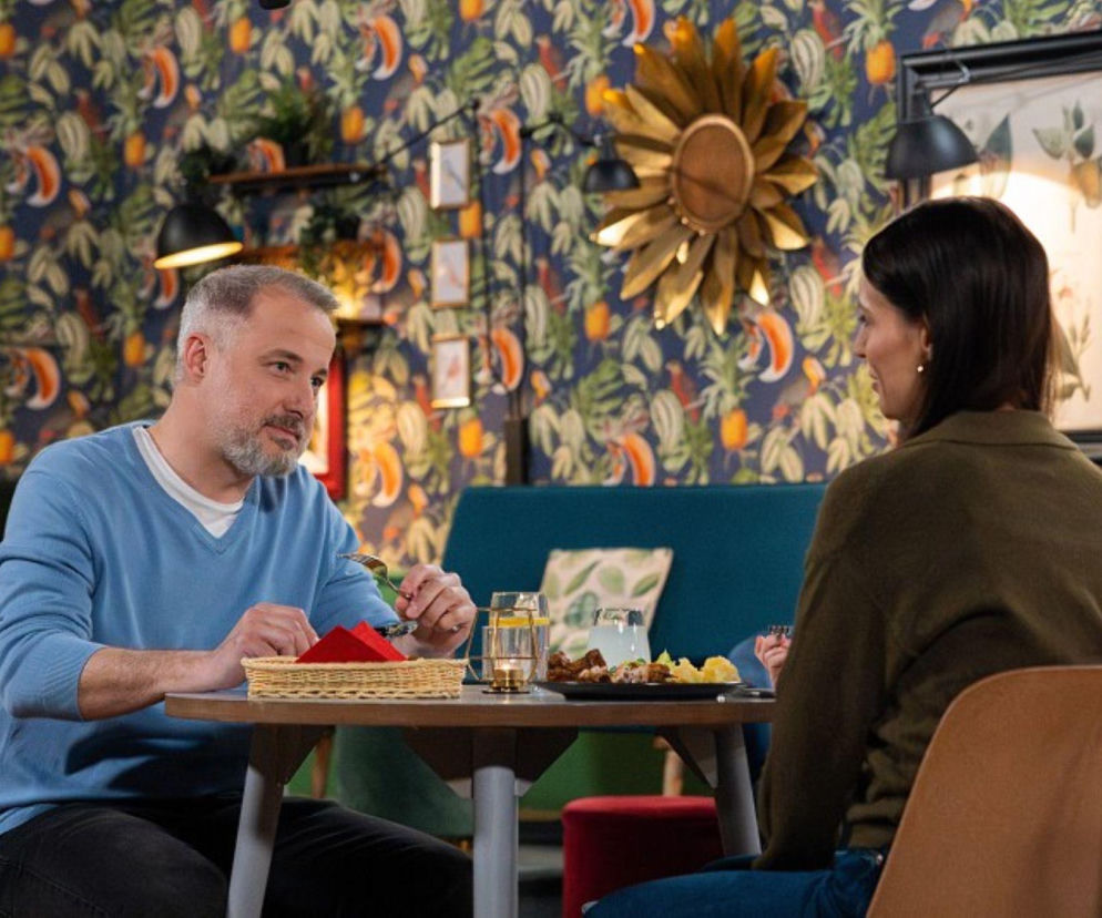 Mężczyzna w błękitnym swetrze i kobieta w zielonej bluzce siedzą przy drewnianym stole w restauracji, jedząc obiad. W tle widać wzorzystą tapetę z motywem liści i egzotycznych ptaków, oraz złote ozdoby ścienne. Zdjęcie ilustruje relację Bartka i Ilony z serialu Pierwsza miłość, o której możesz przeczytać na Super Seriale.