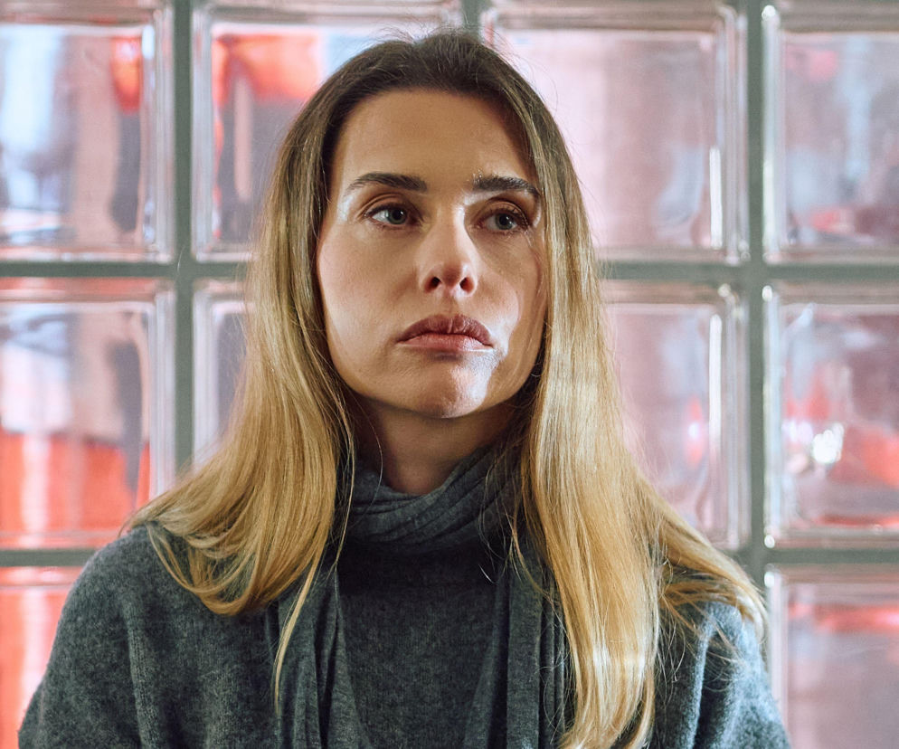 Zbliżenie na Elizę z serialu Na Wspólnej, ze zmartwionym wyrazem twarzy, patrzącą w dal. Jej jasne włosy swobodnie opadają na ramiona. Ubrana jest w szary golf. Tło tworzą rozmyte, przeszklone elementy. Całość oddaje napięcie wokół fabuły Super Seriale.