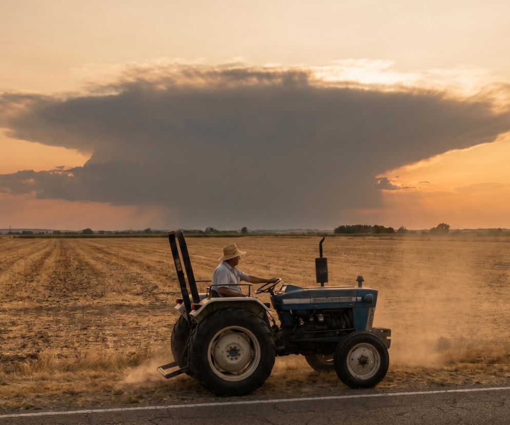 Mężczyzna w słomkowym kapeluszu i jasnej koszuli prowadzi traktor po drodze gruntowej na tle pola i zbliżającej się burzowej chmury, co symbolizuje ostrzeżenia pogodowe, o których przeczytasz na naszym portalu.