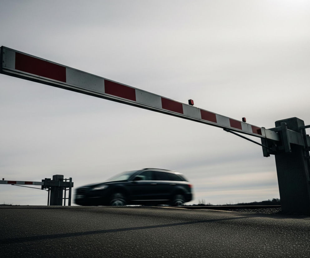 Pozioma szlabanowa zapora kolejowa, pomalowana w naprzemienne biało-czerwone pasy i wyposażona w trzy czerwone światła ostrzegawcze, unosi się ukośnie w górę, zajmując centralną część kadru. Po prawej stronie szlabanu widać ciemną konstrukcję podtrzymującą, a po lewej, w oddali, zarys drugiego, niżej położonego szlabanu. Pod główną zaporą, przez jezdnię, przejeżdża rozmyty, ciemny samochód osobowy, pozostawiając za sobą ślad światła. Tło stanowi jasne, zachmurzone niebo, z jaśniejszym obszarem w prawym górnym rogu.