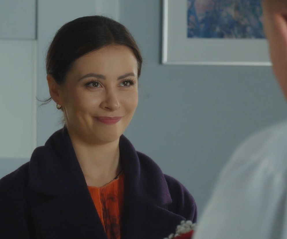 Joanna (Dominika Sakowicz) w fioletowym płaszczu i pomarańczowej bluzce z uśmiechem patrzy na Artura (Robert Moskwa), którego widać z tyłu w jasnej koszuli. Zdjęcie ilustruje scenę zbliżenia Joanny i Artura, gdzie ona wręcza mu bukiet róż. Więcej o ich relacji w M jak miłość przeczytasz na Super Seriale.