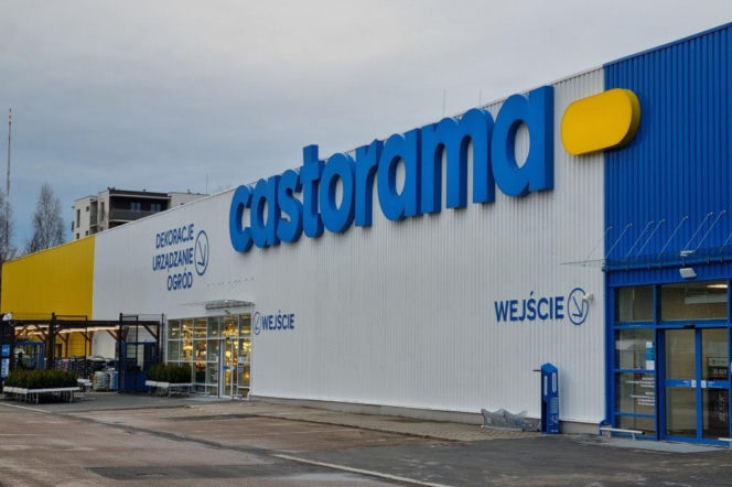 Zewnętrzny widok budynku sklepu Castorama Smart w Ostrowie Wielkopolskim z niebieskim logotypem i białymi ścianami, częściowo z żółtą elewacją. Widoczne wejścia do marketu budowlanego oraz strefa ogrodowa. Więcej o zamknięciu sklepu przeczytasz na Super Biznes.