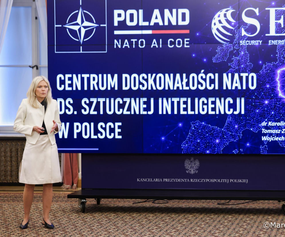 Kobieta w białym kostiumie prezentująca projekt Centrum Doskonałości NATO ds. Sztucznej Inteligencji w Polsce. W tle niebieski ekran z logotypami NATO, POLAND NATO AI COE i SET, przedstawiający mapę Polski jako centrum sieci połączeń. Więcej informacji o tej inicjatywie znajdziesz na naszym portalu.