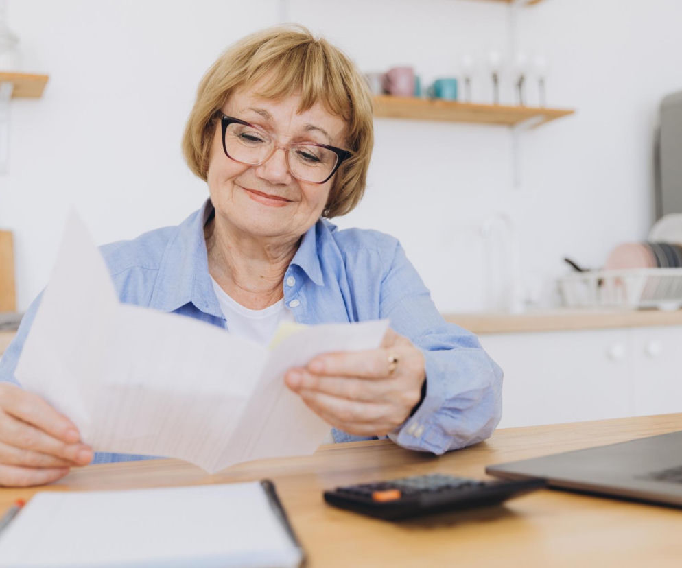 Starsza kobieta w okularach przeglądająca dokumenty przy stole w jasnej kuchni, obok laptop i kalkulator. Artykuły na Super Biznes informują o ulgach PIT-0 dla seniorów.