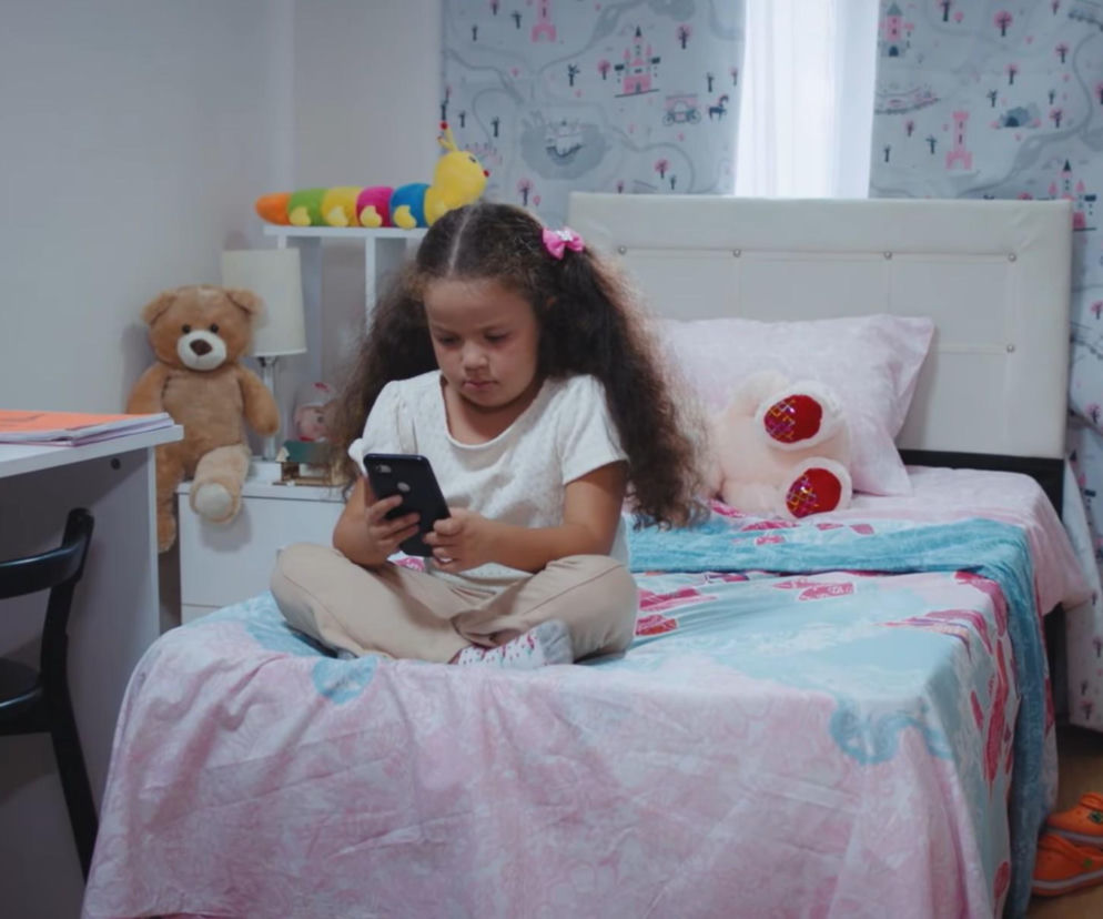 Dziewczynka z ciemnymi, kręconymi włosami i różowymi spinkami, siedząca na łóżku z telefonem w rękach. Na łóżku jest różowa pościel z dziecięcym wzorem, obok pluszowe misie i na półce kolorowa gąsienica. Kontekst dotyczy serialu Dziedzictwo, a więcej o nim znajdziesz na stronie Super Seriale.