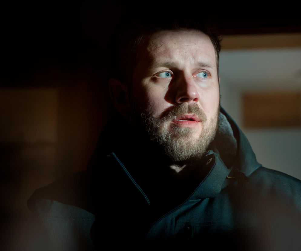 Mężczyzna z brodą i jasnymi oczami, wyraźnie zaskoczony lub zaniepokojony, spogląda w prawą stronę. Na jego twarzy widać cień, co podkreśla dramatyczny nastrój. Całość pasuje do emocjonującego odcinka serialu Uroczysko, dostępnego na Super Seriale.