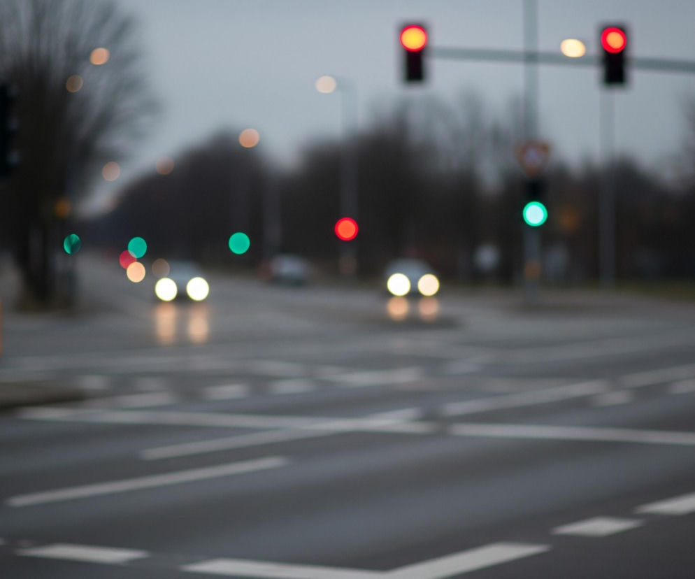 Rozmazany widok nocnego skrzyżowania przedstawia ulicę z białymi liniami pasów i przejść dla pieszych. Na niebie widać dwa czerwone światła sygnalizacji świetlnej po prawej stronie oraz jedno zielone, a po lewej stronie niewyraźne zielone światło. Reflektory samochodów, zarówno z przodu, jak i z tyłu, są widoczne jako rozmyte, jasne punkty świetlne na jezdni. Tło to rozmyte drzewa i lampy uliczne w odcieniach szarości i brązu.