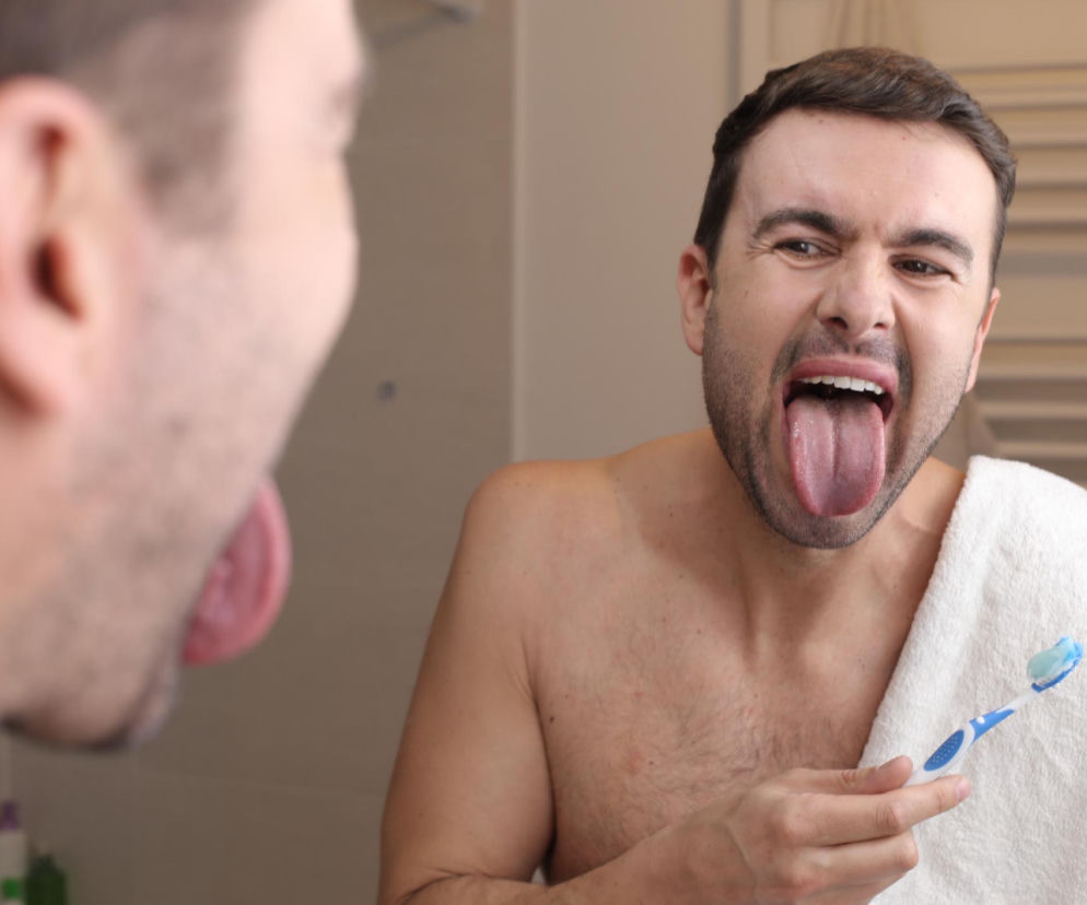 Mężczyzna bez koszulki w lustrze, wyciągający język z widocznym nalotem, trzymający szczoteczkę do zębów z pastą, gotowy do porannej higieny. Artykuł na Poradnik Zdrowie wyjaśnia, co kolor nalotu na języku mówi o stanie jelit i zdrowiu.