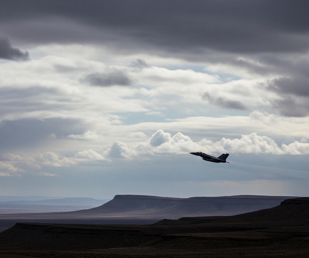Sylwetka ciemnego samolotu odrzutowego z ostrym, spiczastym nosem i charakterystycznymi skrzydłami w kształcie delty unosi się na prawo od środka kadru, pozostawiając za sobą białą smugi kondensacyjną, skierowaną ku górze w lewą stronę. Pod nim rozciąga się ciemny, pustynny krajobraz z płaskimi wierzchowinami wzniesień i horyzontem z warstwami zamglonych gór, od jasnofioletowego po ciemny brąz. Górną część obrazu wypełnia zachmurzone niebo z szarymi, puszystymi chmurami, przeplatanymi jaśniejszymi fragmentami niebieskiego nieba, tworząc dynamiczne tło dla lecącego odrzutowca.