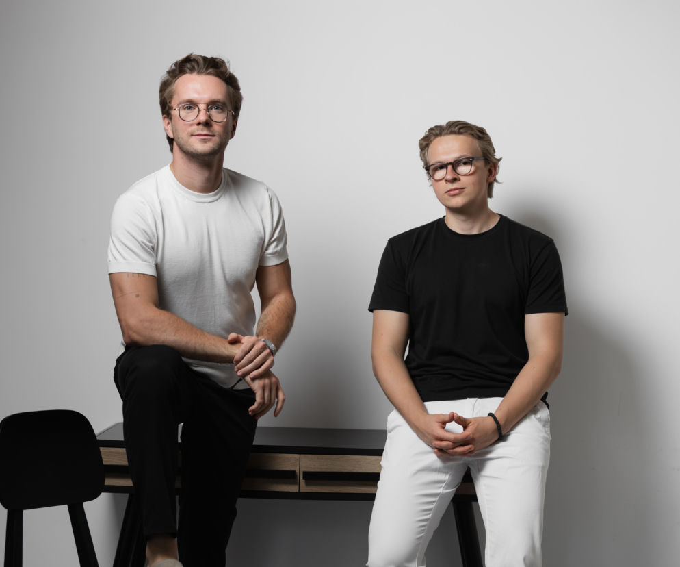 Dwóch młodych mężczyzn w okularach, jeden w białej koszulce i czarnych spodniach siedzący na biurku, drugi w czarnej koszulce i białych spodniach stojący obok. Obaj prezentują się profesjonalnie, co pasuje do tematu artykułu o telemedycynie na Poradnik Zdrowie.