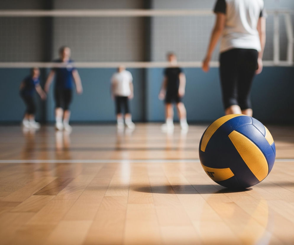 Na pierwszym planie, po prawej stronie leży piłka do siatkówki, częściowo widoczna. Ma ona panele w kolorze granatowym i żółtym. Piłka rzuca wyraźny cień na jasną, drewnianą podłogę sali gimnastycznej. W tle, za piłką i siatką, widać grupę niewyraźnych postaci ubranych w stroje sportowe, grających w siatkówkę.
