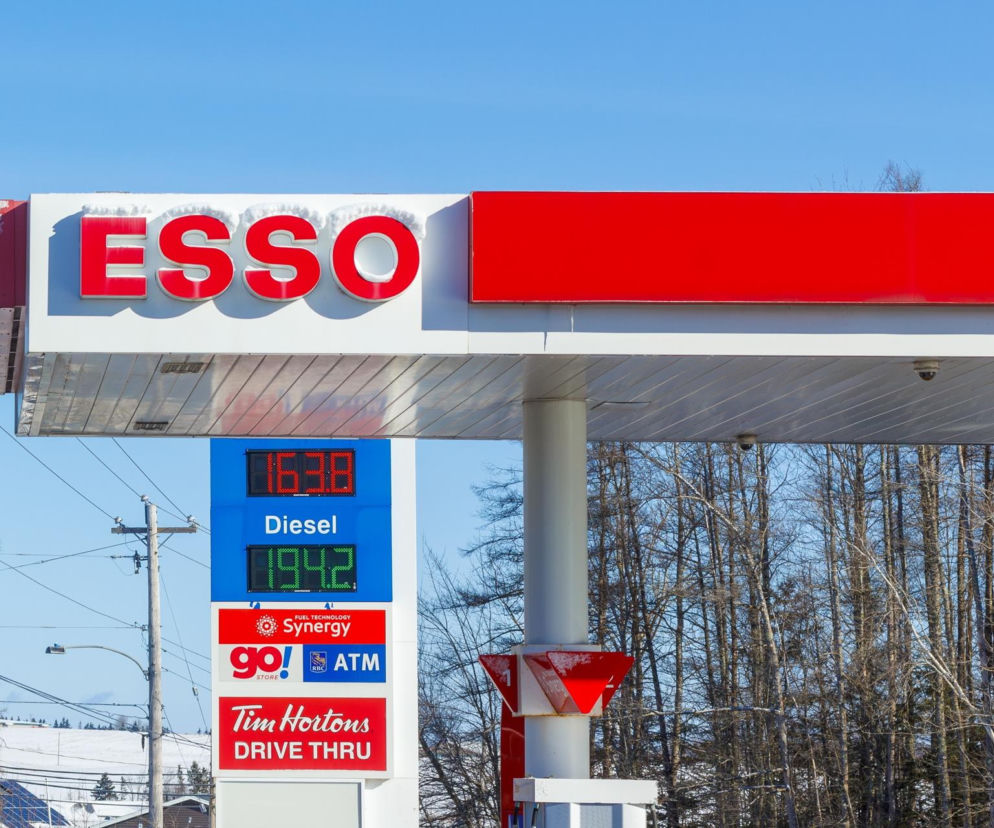 Stacja paliw Esso z tablicą cenową, na której widoczne są ceny oleju napędowego i benzyny, na tle niebieskiego nieba. Po lewej stronie znajduje się słup energetyczny, a w tle drzewa. Aktualne ceny paliw na Super Biznes.