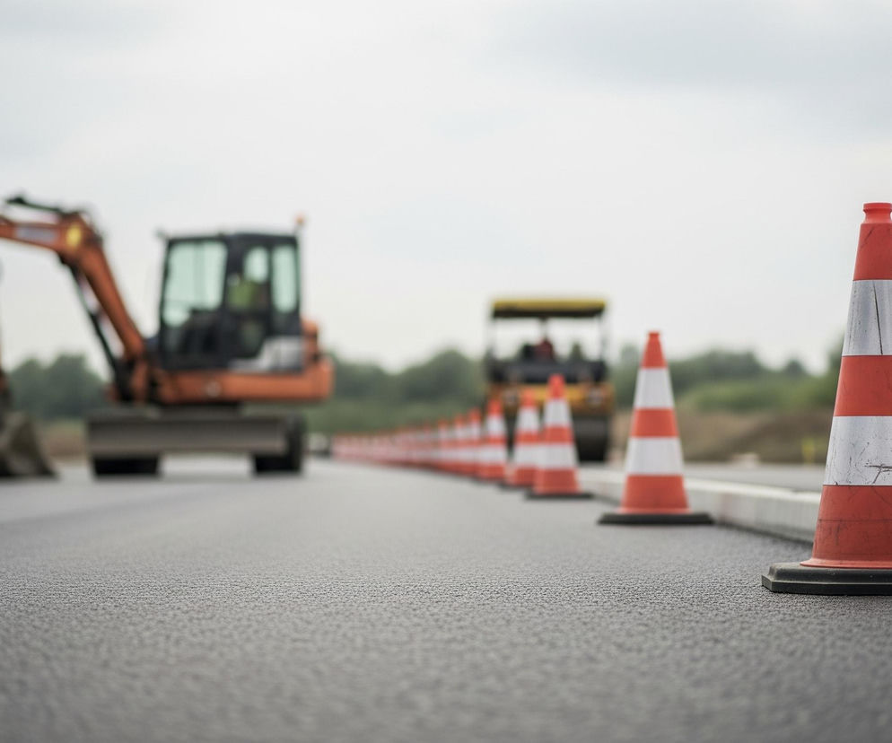 Pionowy, pomarańczowo-biały pachołek drogowy stoi na czarnym asfalcie w prawym dolnym rogu obrazu, blisko białego krawężnika. Za nim, w oddali, wzdłuż nowej nawierzchni, ciągnie się rząd podobnych pachołków, tworząc perspektywiczny zbieg w lewo. W tle, rozmazane, widoczne są pomarańczowe maszyny budowlane, w tym koparka z otwartą łyżką i walec, wszystko pod jasnym, zachmurzonym niebem.