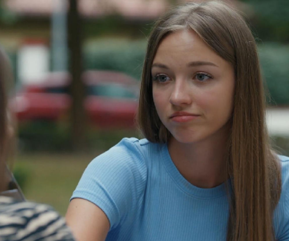 Młoda kobieta, Ewelina (Amelia Listwoń) z serialu „Barwy szczęścia”, z długimi brązowymi włosami i w błękitnej bluzce, patrzy w bok z lekkim niezadowoleniem. W tle widać niewyraźne zarysy drzew i czerwonego samochodu. O sporze Ewy i Eweliny przeczytasz na Super Seriale.