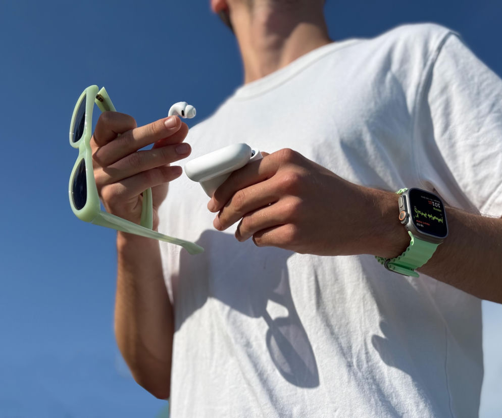 Mężczyzna w białym T-shircie trzyma w dłoniach jasnozielone okulary przeciwsłoneczne i białe słuchawki douszne AirPods Pro 3, wyjęte z etui. Na nadgarstku zielony smartwatch z widocznym pomiarem tętna. Zdjęcie ilustruje recenzję słuchawek Apple, którą można przeczytać na naszym portalu.